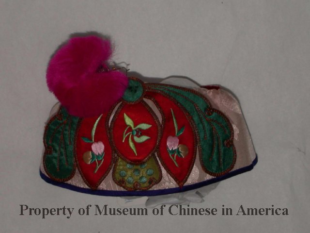 1995.001.003 Pink silk children's headband with colorful lotus flower embroidery at front. Red cotton lining; blue trim at top; hot pink pompom connected to bottom of lotus embroidery by metal wire.  Elastic band at bottom of headband. Courtesy of Lily Hou, Museum of Chinese in America (MOCA) Collection.
粉红色丝绸儿童头带，正面饰有彩色莲花刺绣。红色棉衬里；顶部有蓝色饰边；粉红色绒球通过金属线连接到莲花刺绣的底部。头带底部有松紧带。Lily Hou捐赠，美国华人博物馆 (MOCA) 馆藏。