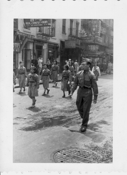 2006.004.447 Man wearing a scouting uniform leading a group of Girl Scouts down a street in New York's Chinatown. The girls are wearing 1940s era Girl Scout uniforms. Courtesy of Marcela Dear, Museum of Chinese in America (MOCA) Collection. 穿着童军制服的男子带领一群女童子军走在纽约唐人街的一条街道上。女孩们穿着 1940 年代的女童子军制服。由陈雪瑛提供，美国华人博物馆 (MOCA) 馆藏 