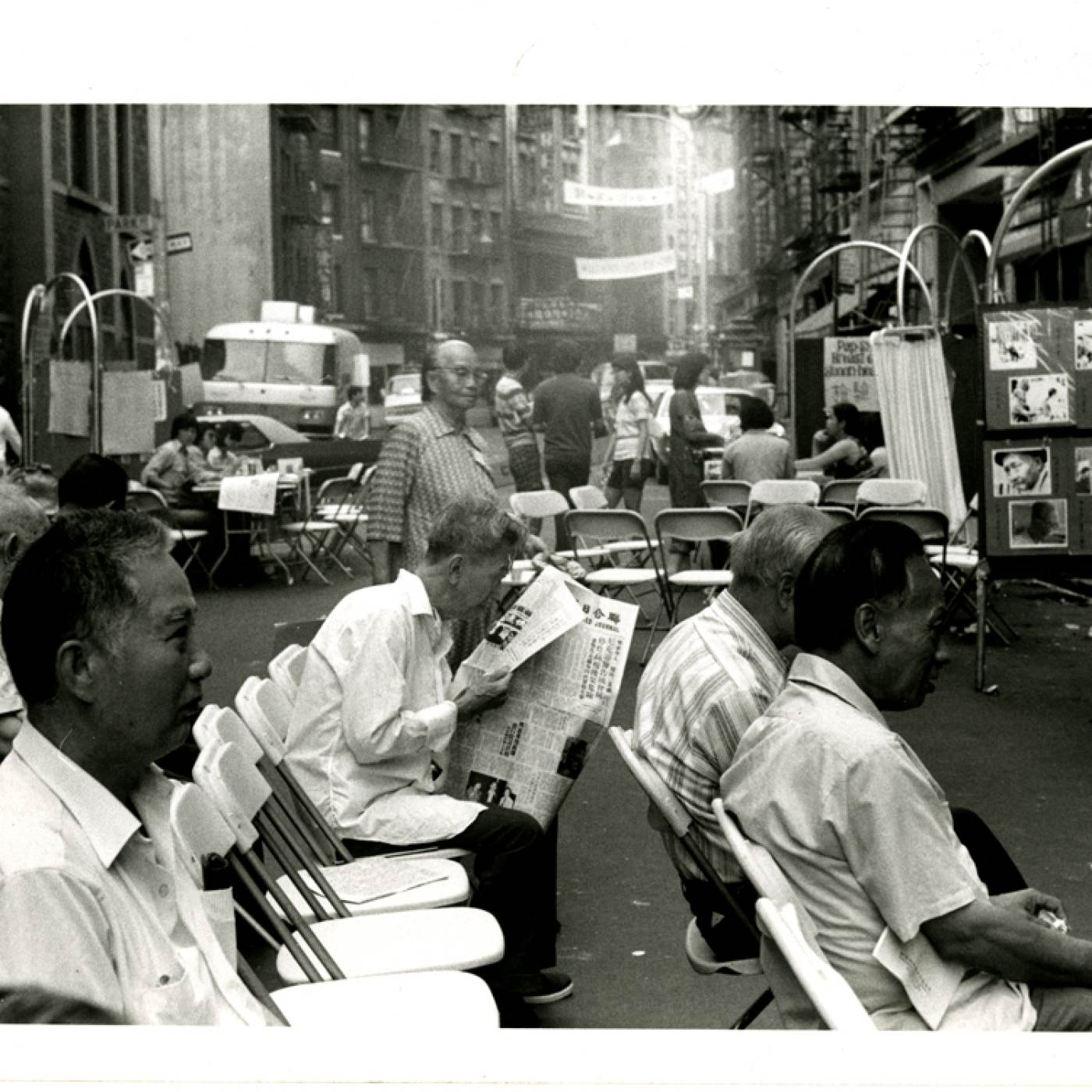 2013.018.011 Photograph taken at the first Chinatown Health Fair on Mott Street at the intersection of Mosco Street in summer 1971. The seated men were waiting for some of the free screening tests (TB Tine, blood pressure, drawing blood, etc.) provided by over 100 medical volunteers and college students trained to administer these simple tests. White folding chairs line the street. Museum of Chinese in America (MOCA) Basement Workshop Collection.  1971年夏天在勿街与莫士高街交叉口处的第一届唐人街健康博览会上拍摄的照片。坐着的人正在等待着一些免费筛查测试（结核病测试、测血压、抽血等），由超过 100 名医疗志愿者和受过训练的大学生来管理这些简单的测试。白色的折叠椅沿街排列。美国华人博物馆（MOCA）地下室工作坊馆藏。