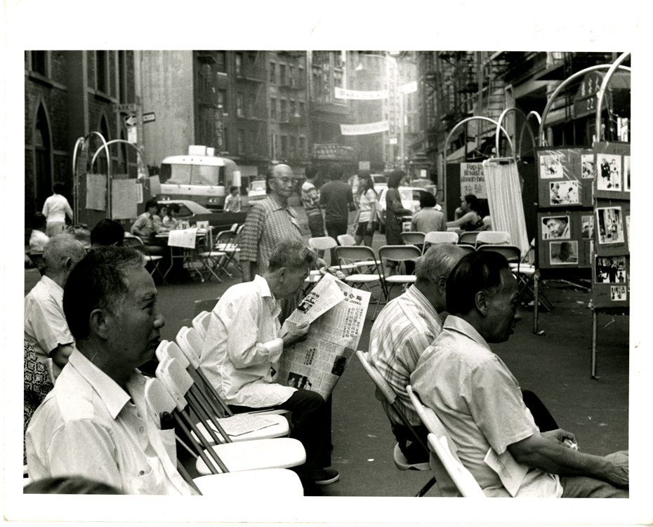 2013.018.011 Photograph taken at the first Chinatown Health Fair on Mott Street at the intersection of Mosco Street in summer 1971. The seated men were waiting for some of the free screening tests (TB Tine, blood pressure, drawing blood, etc.) provided by over 100 medical volunteers and college students trained to administer these simple tests. White folding chairs line the street. Museum of Chinese in America (MOCA) Basement Workshop Collection.  1971年夏天在勿街与莫士高街交叉口处的第一届唐人街健康博览会上拍摄的照片。坐着的人正在等待着一些免费筛查测试（结核病测试、测血压、抽血等），由超过 100 名医疗志愿者和受过训练的大学生来管理这些简单的测试。白色的折叠椅沿街排列。美国华人博物馆（MOCA）地下室工作坊馆藏。